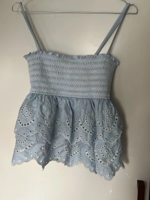 Ljusblå broderad linnetopp - Supersöt ljusblå linnetopp med smala axelband och broderade detaljer. Toppen har smock upptill och volangkant nertill med fina hålbroderier. Perfekt för varma dagar och ger en romantisk vibe till din outfit.