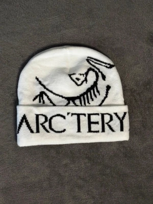 Vit mössa från Arc'teryx - Snygg vit mössa från Arc'teryx med svart logga och text framtill. Klassisk uppvikt kant och mjukt stickat material som håller dig varm. Perfekt för dig som gillar outdoor och streetwear vibes. Vet inte ifall den är äkta eller inte
