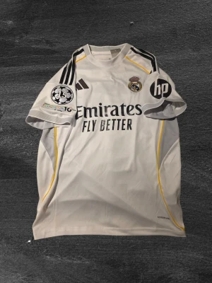 Real Madrid matchtröja Adidas - Vit Real Madrid  VINI.JR.matchtröja med svarta och gula detaljer. Adidas-logga på bröstet, klubbmärke och sponsorer som Emirates och HP. (AEROREADY).