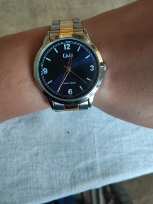 Q&Q klocka med blå urtavla - Stilren klocka från Q&Q med blå urtavla och tydliga vita siffror. Armbandet är i silverfärgad metall med en gulddetalj i mitten. Klassisk design som passar till många stilar. Vattentålig och med justerbart metallarmband.