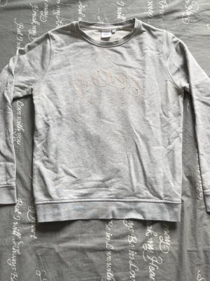 Grå sweatshirt från Hugo Boss - Snygg grå sweatshirt från Hugo Boss med diskret logga i beige och vita detaljer framtill. Tröjan har rund halsringning, långa ärmar och mjukt material som känns skönt mot huden. Perfekt för en avslappnad och stilren look.
