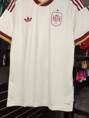 Vit Spanien Adidas fotbollströja - Snygg vit fotbollströja från Adidas med Spaniens landslagsemblem på bröstet. Tröjan har vinröda och gula detaljer vid krage, ärmslut och axlar samt Adidas-logga. Tillverkad i lätt och ventilerande material (Climacool). Perfekt för träning eller match.