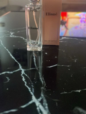 Elinné parfym från NG - Elinné parfym från NG i en elegant, genomskinlig glasflaska med svart text. Flaskan har en stilren de och modern design. Förpackningen är beige med svart text och ger ett exklusivt intryck. Perfekt för dig som vill ha en fräsch och lyxig doft.