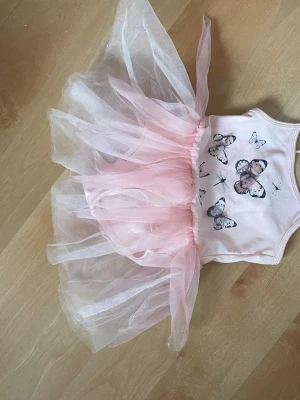 Rosa tyllklänning med fjärilar storlek 92 - Supersöt kort rosa klänning med tyllkjol och fjärilstryck framtill. Klänningen är ärmlös och har flera lager ljusrosa tyll som ger en fluffig och lekfull look. Perfekt för dig som gillar gulliga detaljer och mjuka pastellfärger.