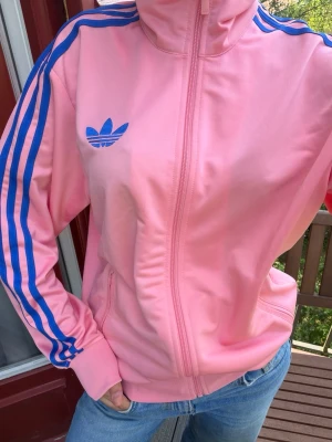 Rosa Adidas track jacket med blå ränder - Säljer en snygg rosa Adidas track jacket med blå klassiska ränder längs ärmarna. Jackan har dragkedja framtill, hög krage och muddar vid ärmsluten