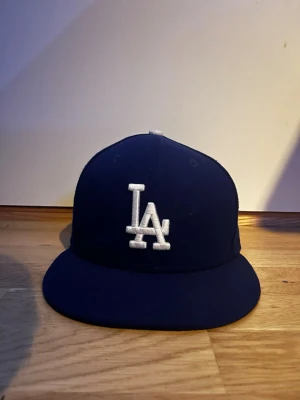 Blå LA Dodgers keps från New Era - Mörkblå keps med rak skärm och broderad LA-logga i vitt framtill. Klassisk modell från New Era, perfekt för dig som gillar sportig streetstyle. Kepsen har en strukturerad form och ventilation i ovandelen.