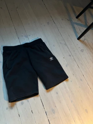 Svarta shorts från Adidas - Svarta shorts från Adidas med klassisk logga på vänster ben. Mjuk resår i midjan och två sidofickor. Perfekta för chill dagar eller träning. Tillverkade i bomull för skön känsla och enkel stil. 