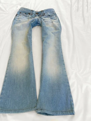 Ljusblå bootcut jeans från Nudie - Säljer ett par ljusblå bootcut jeans från Nudie Jeans med snygg slitning på låren och klassiska fem fickor. Jeansen har låg midja och vida ben, perfekt för en avslappnad och trendig look.