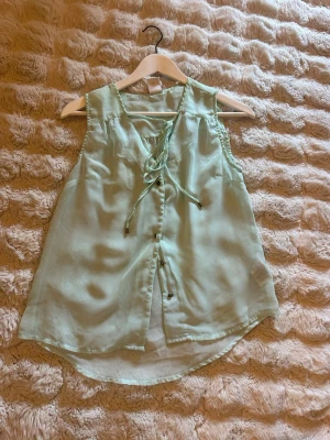 Mintgrön ärmlös blus från Vero Moda - Säljer en mintgrön, ärmlös blus från Vero Moda. Blusen har knytning vid halsen, knappar framtill och är tillverkad i ett tunt, luftigt material som passar perfekt till varma dagar. Lätt rundad nederkant för en avslappnad look. Lösa trådar och pyttesmå hål förekommer 