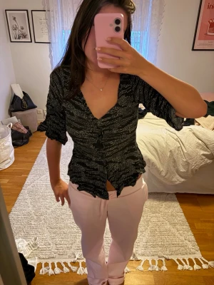 Svart v-ringad blus med volang - Trendig svart blus med vitt mönster och v-ringning. Blusen har volangkant nertill, knappar framtill och trekvartsärm med lätt puff. Sitter figurnära och ger en snygg siluett. Perfekt för dig som vill ha en cool och feminin topp.