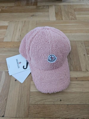 Rosa teddykeps från Moncler - Säljer en fluffig rosa keps från Moncler i teddy-material. Kepsen har böjd skärm och ett broderat Moncler-märke framtill. Perfekt för dig som vill ha en unik och trendig accessoar som sticker ut i mängden.