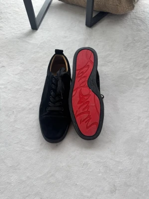 Svarta Christian Louboutin sneakers - Snygga svarta sneakers från Christian Louboutin med klassisk röd sula och svart mocka på ovandelen. Skorna har svarta skosnören och låg profil. Perfekta för dig som vill ha något exklusivt och stilrent med en unik touch. Litet släpp i en söm på ena skon.