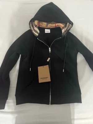 Svart hoodie från Burberry - TRÖJAN ÄR HELT NY OCH OANVÄND.                                            Säljer för att den inte passar och är för liten.                                                           Svart hoodie från Burberry med dragkedja och klassiskt rutigt foder i huvan. Snygga snören med metalländar och två fickor framtill. Perfekt för dig som vill ha en stilren och lyxig look. Materialet är mjukt och bekvämt. Priset kan diskuteras.