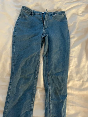 Blå raka jeans med hög midja - Säljer ett par klassiska blå jeans med raka ben och hög midja. Jeansen har dragkedja och knapp framtill samt fickor både fram och bak. Materialet är denim och färgen är en tidlös blå nyans. Perfekta för dig som gillar en enkel och clean look.