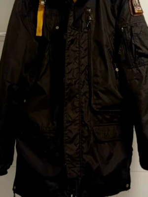 Svart parkas från Parajumpers (Sommarpris) - Säljer en svart parkas från Parajumpers Masterpiece Series. Jackan har flera fickor, dragkedjor och en gul detalj vid kragen. Perfekt för kalla dagar med sin slitstarka och fodrade design. En defekt dragare/knapp på sidan, kostar 150kr hos skräddaren.