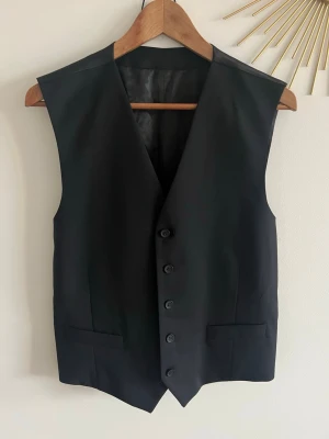 Klassisk svart kostymväst - Stilren svart kostymväst med fyra knappar framtill och två fickor. Västen har slät framsida och blank baksida med justerbart band. Perfekt för att lyfta en outfit med en elegant touch. Passar till skjorta eller t-shirt för en dressad look.