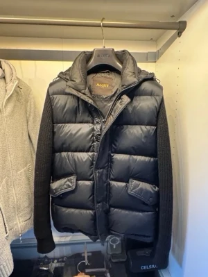 Svart dunväst/hubridjacka från MooRER - Snygg svart dunväst från MooRER med avtagbara stickade ärmar och huva. Västen har två stora fickor framtill med lock och knappar, samt dragkedja och knappstängning. Storlek 48it men passar S/M