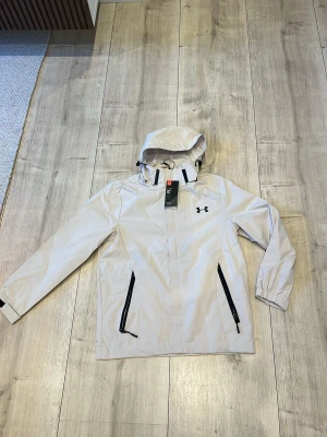 Ljusgrå regnjacka från Under Armour - Snygg ljusgrå regnjacka från Under Armour med svart logga på bröstet. Jackan har huva med dragsko, svarta dragkedjor på fickorna och justerbara ärmslut. Perfekt för regniga dagar och outdoor-aktiviteter.