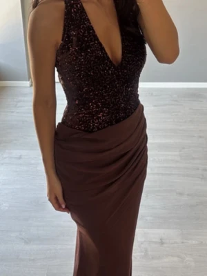 Klänning från Bella Barnett   - Snygg långklänning i brun nyans med glittrig överdel och djup v-ringning. Klänningen har ärmlös design och figurnära passform med draperad kjol. Perfekt för dig som vill sticka ut med elegans och stil.