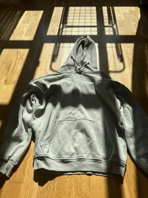 Ljusgrå basic hoodie med huva - En stilren ljusgrå hoodie med klassisk huva och känguruficka framtill. Tröjan har ribbade muddar vid ärmslut och nederkant samt en avslappnad passform. Perfekt för en chill och enkel look. Materialet ser ut att vara mjuk bomullsblandning.