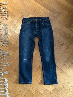Nudie jeans  - Säljer ett par klassiska blå jeans från Nudie Jeans med snyggt slitna detaljer och kontrastsömmar. Jeansen har fem fickor, raka ben och en cool broderad design på bakfickorna. Perfekt för dig som gillar en avslappnad och trendig look.