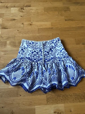 Vit och blå volangkjol från Zara - Supersnygg kort kjol från Zara i vitt och blått med volangkant och ett unikt mönster. Kjolen har hög midja och dragkedja bak. Perfekt för dig som vill sticka ut med en cool och somrig look!
