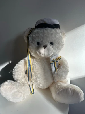 Vit studentnalle med mössa och band - Stor söt nallebjörn med studentmössa och blågula band. Stått mycket väl förvarad sedan min student. Någon kanske hade blivit glad för denna söta nallebjörn nu till studenten. 