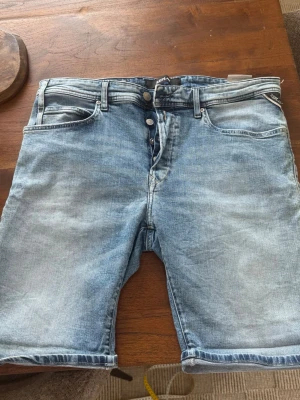 Blå jeansshorts från Replay - Snygga blå jeansshorts från Replay med klassisk femficksmodell och knappar i gylfen. Shortsen har en ljus tvätt och subtila slitningar för en avslappnad look. Perfekta för varma dagar och enkel att matcha med olika stilar.
