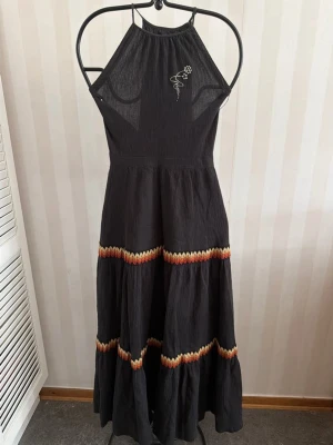 Svart vintage  långklänning med broderi - Svart långklänning med halterneck och broderad blomdetalj på bröstet. Klänningen har volangdetaljer och färgglada vågiga band i rött, gult och orange. Tillverkad i ett lätt, skrynkligt bomullstyg och har ett matchande bälte i midjan.