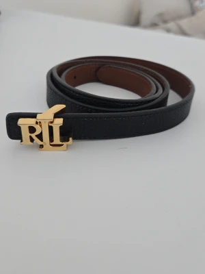 Svart skärp från Ralph Lauren - Snyggt svart skärp från Ralph Lauren med guldfärgat RLL-spänne. Bältet är smalt och har en slät yta med brun insida. Har gjort egengjorda hål i skärpet, använt en del. Köpte den år 2020. 