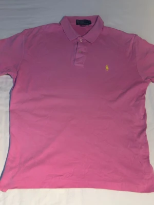 Rosa pikétröja från Ralph Lauren - Säljer en klassisk rosa pikétröja från Ralph Lauren med gul broderad logga på bröstet. Tröjan har krage, knappar framtill och är kortärmad. Perfekt för dig som gillar stilrena och tidlösa plagg.