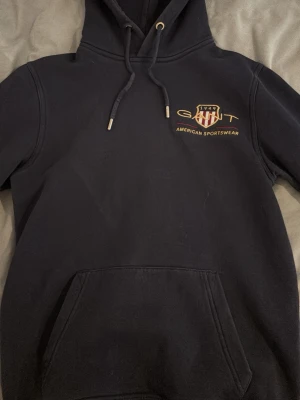 Svart hoodie från GANT med logga - Snygg svart hoodie från GANT med broderad logga och texten 'American Sportswear' på bröstet. Klassisk känguruficka framtill, justerbar huva med dragsko och mjukt material som känns skönt mot huden. Perfekt för en avslappnad och sportig stil.