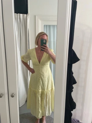 Ljusgul långklänning med broderade detaljer - Ljusgul långklänning med v-ringning och puffärmar. Klänningen har broderade blommönster och volang nedtill, samt fina spetsdetaljer längs sömmarna. Perfekt för sommarens alla tillfällen.