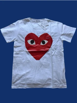 Vit Comme des Garçons Play t-shirt - Vit t-shirt från Comme des Garçons Play med det ikoniska röda hjärtat med ögon tryckt på bröstet. Klassisk rund hals och korta ärmar. Tillverkad i mjuk bomull, perfekt för en avslappnad och trendig stil.