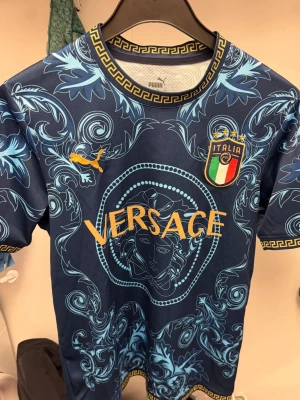 Puma t-shirt Italien - Unik blå t-shirt med Puma-logga samt Italiens landslagsemblem. T-shirten har korta ärmar, detaljerat barockinspirerat mönster i ljusblått och guldiga tryck. Rund halsringning med grekiskt mönster. Perfekt för dig som gillar statement-plagg.