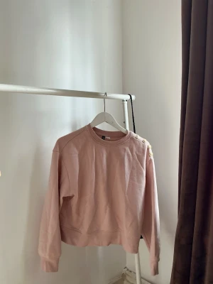 Rosa sweatshirt med knappar på axeln - Säljer en ljusrosa sweatshirt från H&M med rund halsringning och dekorativa guldknappar på ena axeln. Tröjan har en avslappnad passform och ribbade muddar vid ärmslut och nederkant. Perfekt för en chill och stilren look.