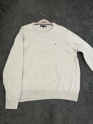 Ljusgrå tröja från Tommy Hilfiger - Ljusgrå rundhalsad tröja från Tommy Hilfiger med diskret broderad logga på bröstet. Tröjan har ribbade muddar vid ärmslut och nederkant. Perfekt för en stilren och enkel look.