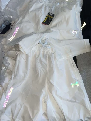 Vita shorts från Under Armour - Snygga vita shorts från Under Armour med elastisk midja och reflekterande logga på benet. Shortsen har en sportig look och är perfekta för träning eller chill. Materialet känns lätt och bekvämt, och det finns praktiska fickor på sidorna.