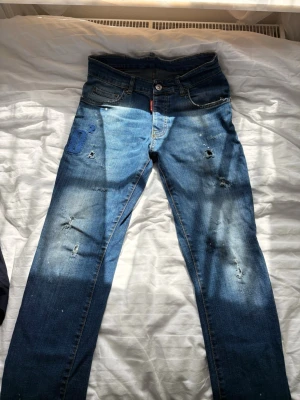  Dsq2 Blå slitna jeans med hål - Säljer ett par blå jeans med slitningar och hål för en trendig look. Jeansen har klassisk femficksdesign, normal midja och smal passform. Perfekta för dig som gillar en avslappnad och edgy stil. Passar från 174-180 