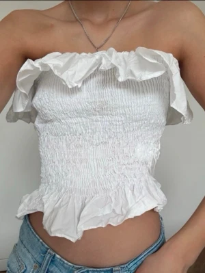 Vit bandeau tubtopp med volang - Säljer en vit bandeau tubtopp med smockad passform och volangdetaljer upptill och nedtill. Toppen är ärmlös och har en luftig, lätt känsla som passar perfekt till jeans eller kjol. Materialet är bomull och toppen har en feminin och trendig look.