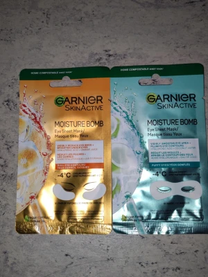 Garnier Moisture Bomb ögonmasker - Två Garnier SkinActive Moisture Bomb Eye Sheet Mask i olika varianter. En har guldigt förpackning med apelsinjuice och den andra är turkos med kokosvatten och hyaluronsyra. Maskerna är veganska och avsedda för att återfukta och reducera svullnad runt ögonen.