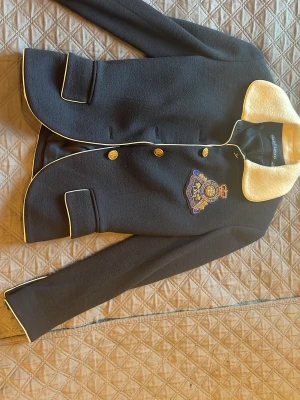 Mörkblå kavaj från Ralph Lauren - Stilren mörkblå kavaj från Ralph Lauren med kontrasterande ljus krage och vita passpoaler. Dubbelknäppt med guldknappar och broderad patch på bröstet. Tillverkad i ullmix med slät insida. Perfekt för en preppy look.
