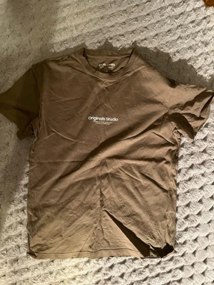 Brun t-shirt från Jack & Jones - Snygg  t-shirt från Jack & Jones Originals Studio med vit text på bröstet. Klassisk passform med rund hals och korta ärmar. Perfekt för en avslappnad stil och enkel att matcha med jeans eller shorts. Hör av dig vid frågor! 