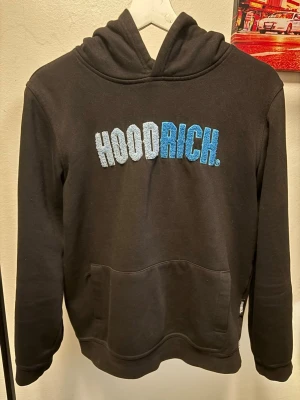 Svart hoodie från Hoodrich 13-15y/ 168 - Svart hoodie från Hoodrich med broderad text i ljusblått och blått på bröstet. Klassisk känguruficka framtill och huva. Perfekt för en streetwear-look och riktigt skön att ha på sig. Passar dig som gillar stilrena och coola plagg. Strl 13-15 men passar även som xs/s