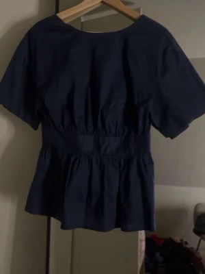 Mörkblå blus med knytskärp - Snygg mörkblå blus med korta ärmar och markerad midja med knytskärp framtill. Blusen har en lätt v-ringning och volang nedtill för en trendig siluett. Perfekt att styla med jeans eller kjol för en cool look.