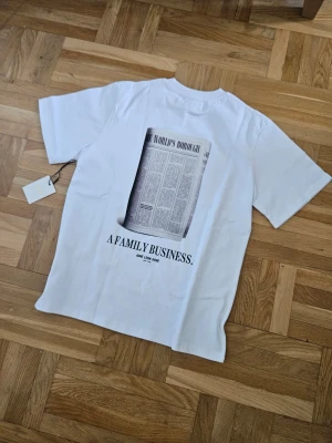 Vit t-shirt Aimé Leon Dore print - Vit t-shirt från Aimé Leon Dore med tryck av en tidningssida och texten 'A FAMILY BUSINESS.' på ryggen. Klassisk passform och rund hals. Perfekt för dig som gillar streetwear och stilrena prints.