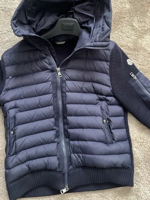 Mörkblå pufferjacka från Moncler - Snygg mörkblå pufferjacka från Moncler med stickade ärmar och vadderad kropp. Jackan har huva, två framfickor med tryckknappar och en ärmficka med dragkedja. Moncler-logga på ärmen och ribbade muddar för extra komfort. Säljer denna eftersom att jag har knappt använt den (pris kan självklart diskuteras.) 