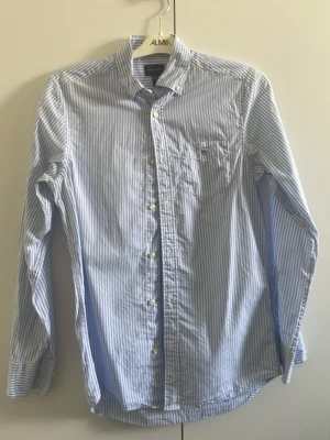 Blårandig skjorta från Gant XS - Klassisk blårandig skjorta från Gant i storlek XS. Skjortan har långa ärmar, knappar framtill och en bröstficka med liten logga. Tillverkad i mjuk bomull och har en tidlös, stilren look som passar perfekt till jeans eller chinos.