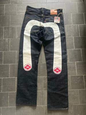 Evisu retro jeans med vit målning, strl 32 ALDRIG ANVÄNDA - Säljer dessa supersällsynta par EVISU-jeans från tidigt 2000-tal. Aldrig använda, private stock, och orginallappen sitter kvar på jeansen. Nypris på deras hemsida för liknande jeans är uppemot nästan 4000kr, du hittar ingen bättre deal för ett par oanvända retrojeans än dessa.😍Perfekt för dig som vill ha ett klädesplagg som är sällsynt, lyxigt, nytt och ingen annan kan ha. De är ganska långa men detta går att åtgärdas enkelt genom att vika upp dem eller att ev.sy. Pris är ej hugget i sten, skriv