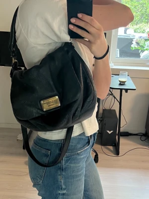 Svart Marc Jacobs handväska - Säljer en svart handväska från Marc by Marc Jacobs i mjukt, strukturerat läder. Väskan har ett lock med guldfärgad metallplatta framtill och både axelrem och handtag. Perfekt för dig som gillar klassisk men cool stil.